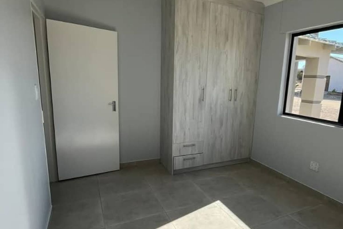 A bright, empty bedroom featuring neutral tiled flooring, a modern built-in wardrobe with wood-grain finish, and clean, light-grey painted walls.