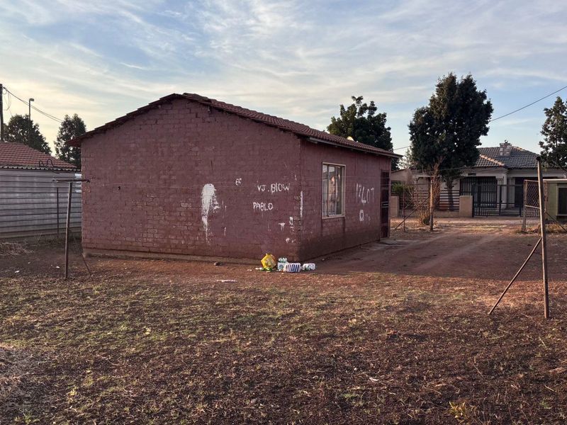 A view of the rear exterior of a brick house featuring a tiled roof and metal window frames, situated on a large unmaintained plot.