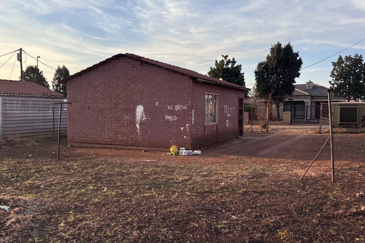 A view of the rear exterior of a brick house featuring a tiled roof and metal window frames, situated on a large unmaintained plot.