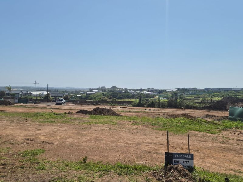 A vacant residential plot of land featuring a for sale sign, situated in a developing estate with surrounding landscape views.