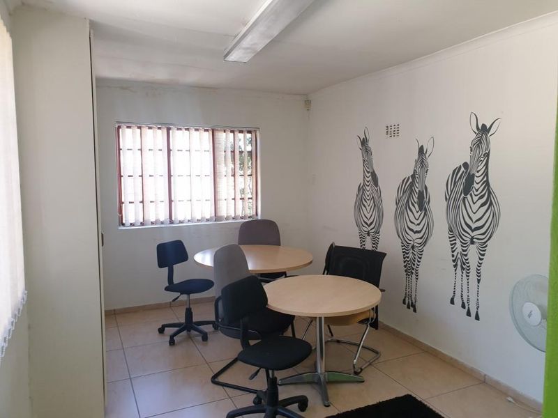 A functional office space featuring tiled flooring, vertical window blinds, and a ceiling-mounted light fixture, currently utilized as a small meeting area.
