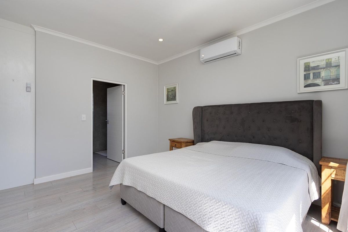 A well-maintained bedroom featuring light-toned laminate flooring, recessed ceiling lighting, and a wall-mounted air conditioning unit, with an adjoining en-suite bathroom.