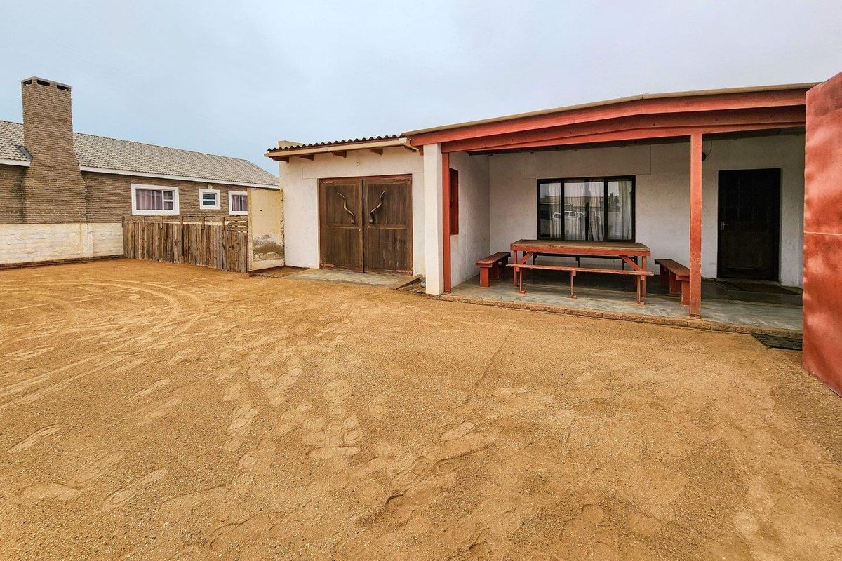 Rear exterior view featuring a covered patio area, timber double doors, and a large open sandy yard space.