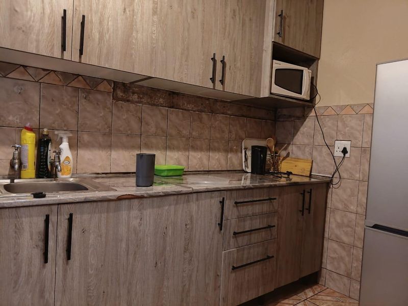 A functional kitchen featuring wood-look laminate cabinetry, a tiled backsplash, and a stainless steel sink, showing signs of wear on the countertop and wall.