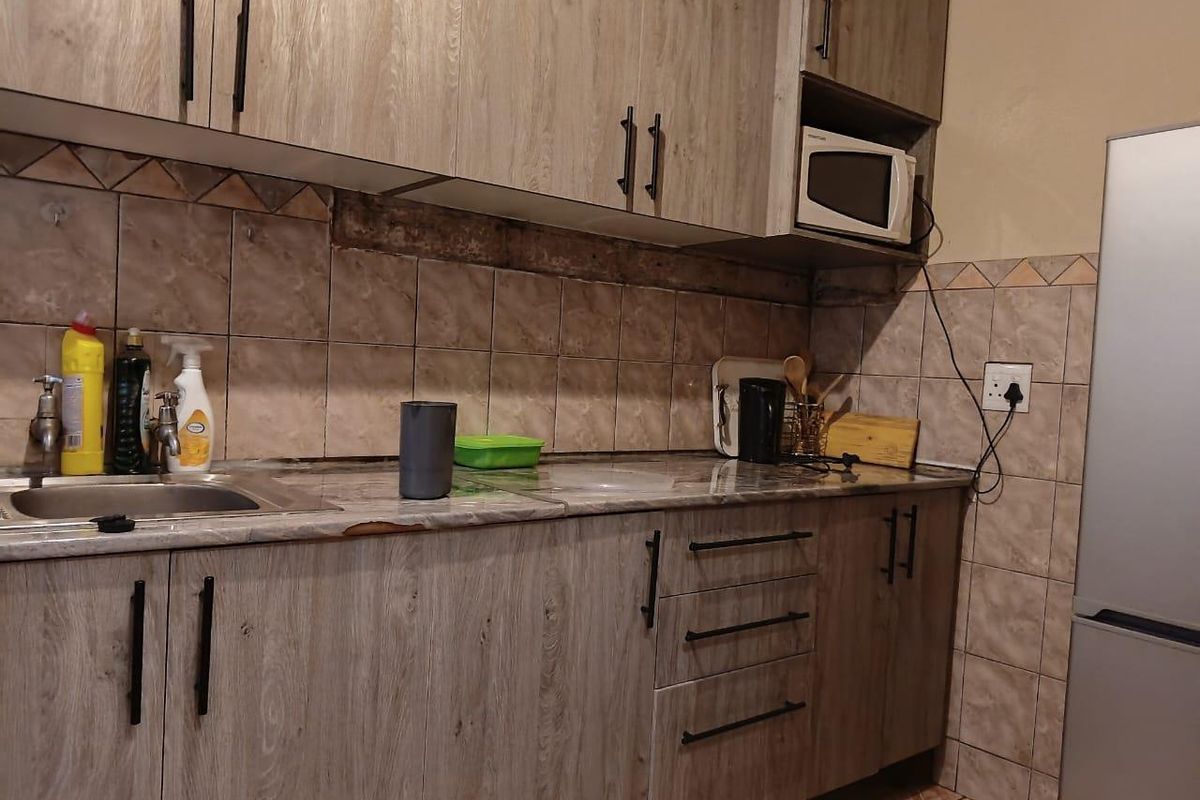 A functional kitchen featuring wood-look laminate cabinetry, a tiled backsplash, and a stainless steel sink, showing signs of wear on the countertop and wall.