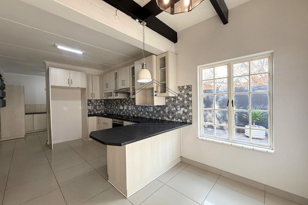 A modern kitchen featuring light wood cabinetry, dark countertops, a breakfast bar, patterned tile backsplash, and exposed ceiling beams.
