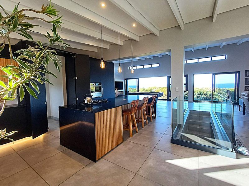 Modern open-plan kitchen and living area featuring a sleek island, exposed beam ceilings, and large sliding doors with scenic coastal views.