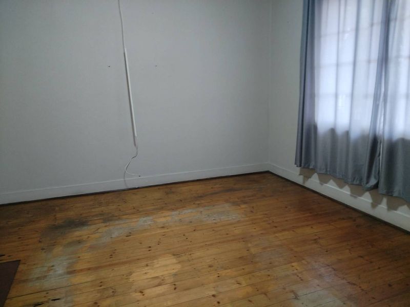An empty bedroom featuring original timber flooring and a window with curtains, requiring cosmetic updates to the floor and wall finishes.