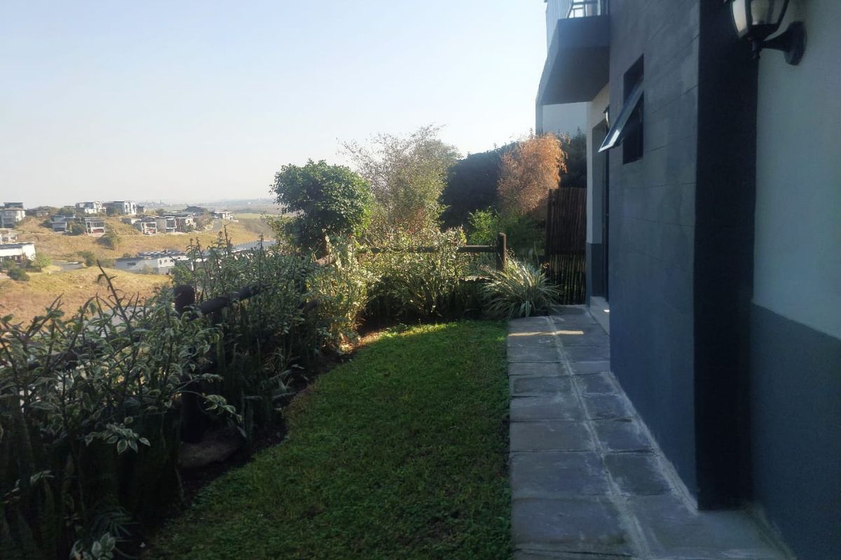 A rear exterior view featuring a stone paved walkway, a small lawn area, and a scenic view of the surrounding estate.