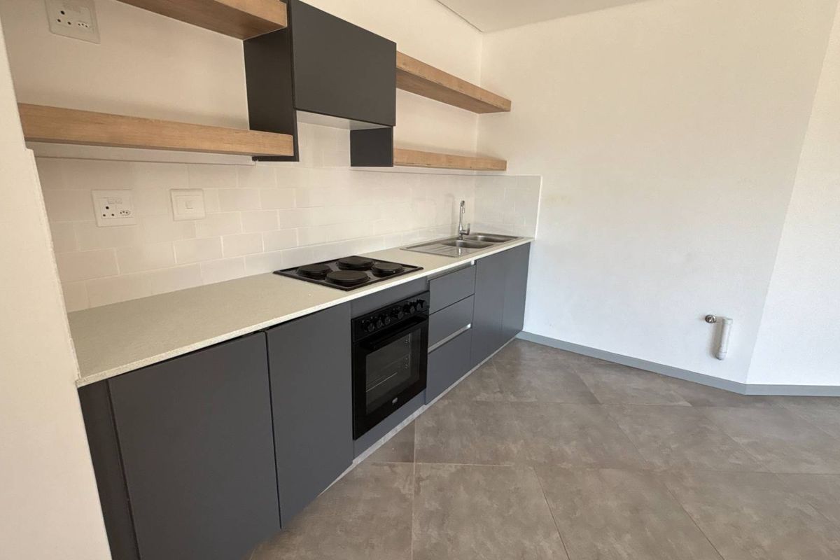 Modern kitchen featuring dark grey cabinetry, light wood floating shelves, built-in oven, electric hob, and subway tile backsplash with neutral floor tiling.