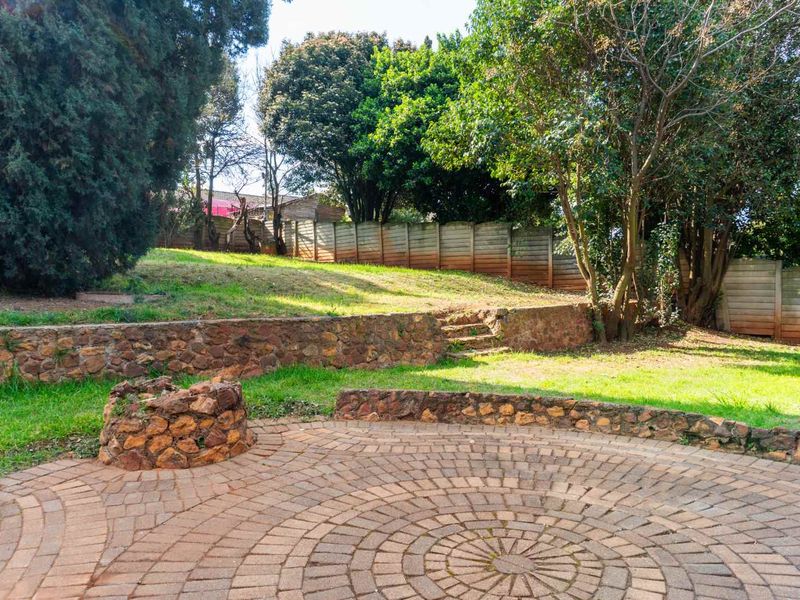 A spacious garden featuring a circular paved patio, stone retaining walls, and mature trees, offering a private outdoor space with established landscaping.