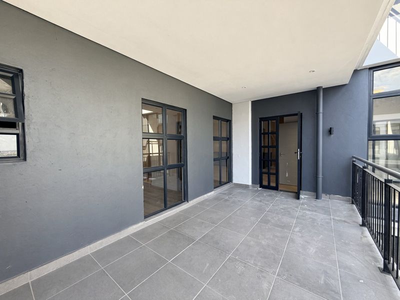 A modern, spacious covered balcony featuring grey tiled flooring, dark grey exterior walls, and black aluminium framed windows and doors with a metal balustrade.