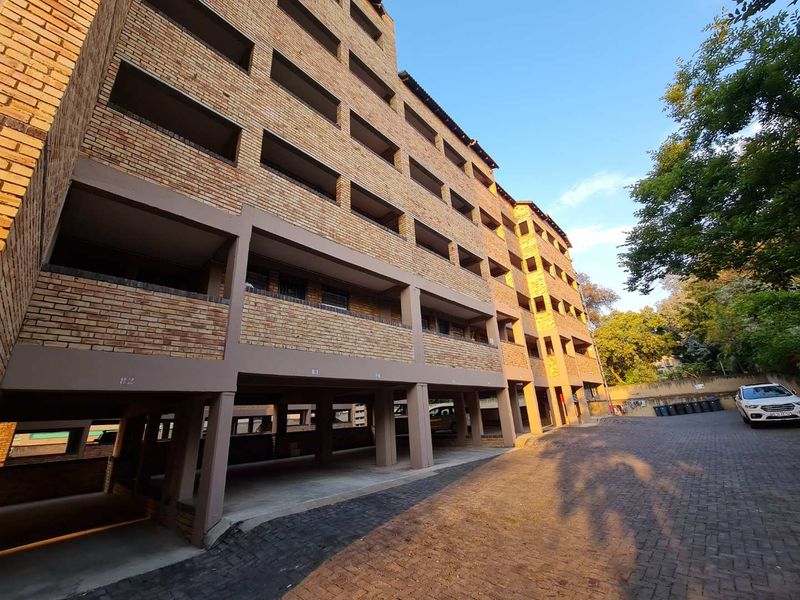 Exterior view of a multi-story apartment building featuring a face brick facade, covered ground-level parking bays, and a paved driveway.