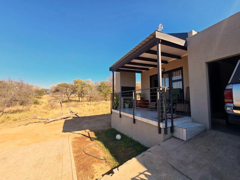 A covered outdoor patio area featuring tiled flooring, a built-in braai, and metal railings, overlooking the surrounding natural landscape.