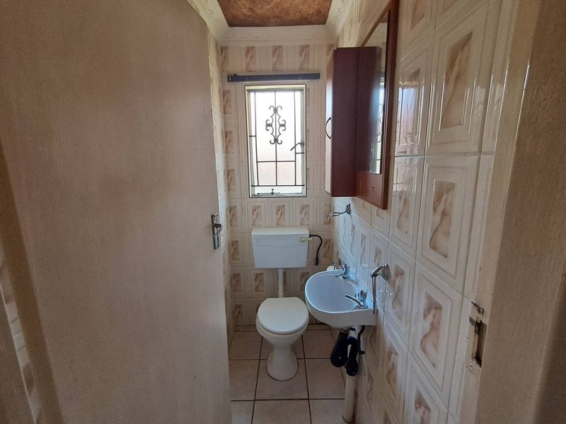 A compact, dated bathroom featuring ceramic wall and floor tiles, a wall-mounted basin, and a standard toilet with a security-gated window.