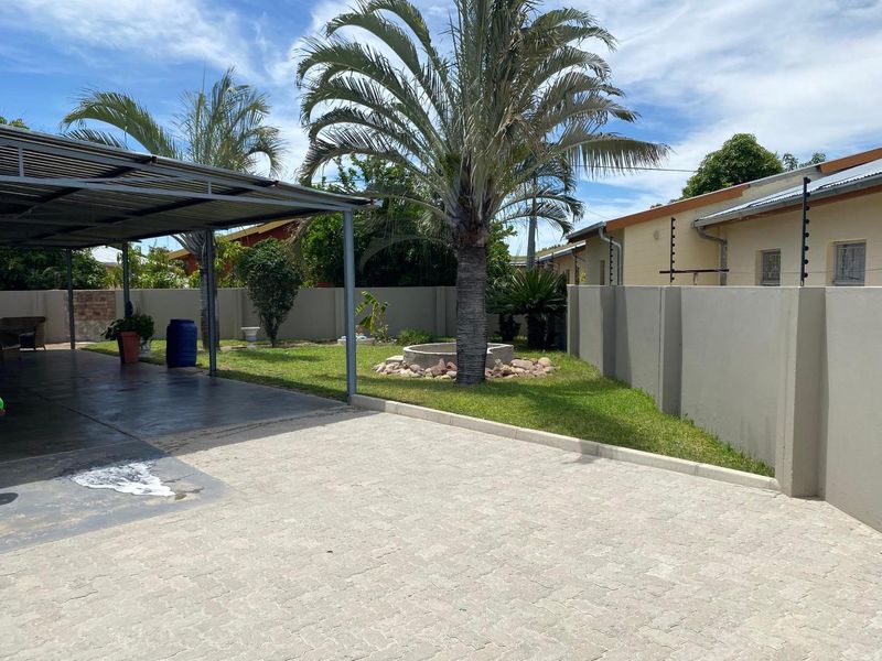 A spacious rear yard featuring a paved driveway, a covered carport, a well-maintained lawn, and secure perimeter walling with electric fencing.