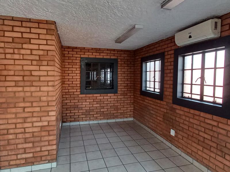 An empty office space featuring exposed brick walls, tiled flooring, security-barred windows, and a wall-mounted air conditioning unit.