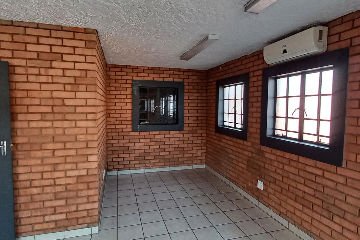 An empty office space featuring exposed brick walls, tiled flooring, security-barred windows, and a wall-mounted air conditioning unit.