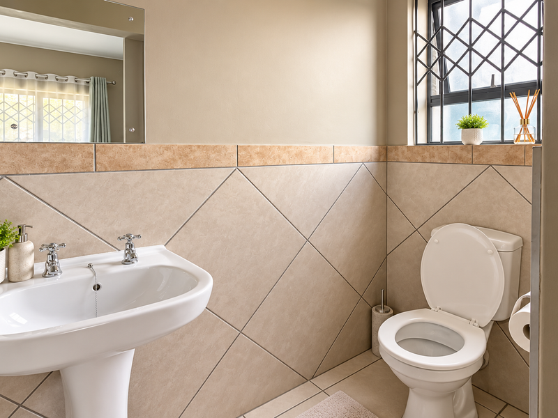 A clean and well-maintained bathroom featuring a pedestal washbasin, ceramic wall and floor tiling, and a window with security bars.
