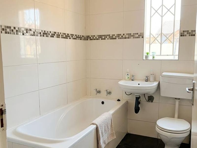 A functional bathroom featuring a built-in bathtub, wall-mounted basin, toilet, and neutral wall tiling with a decorative mosaic border.