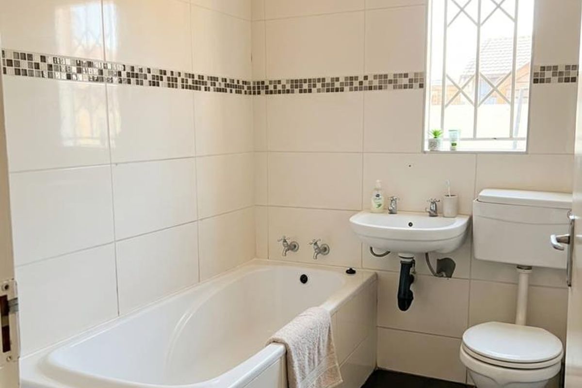 A functional bathroom featuring a built-in bathtub, wall-mounted basin, toilet, and neutral wall tiling with a decorative mosaic border.