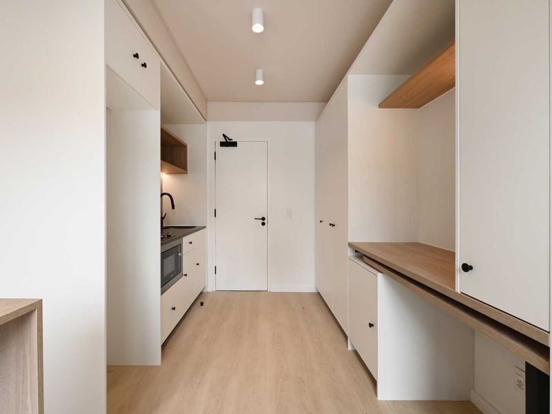 A modern, minimalist kitchen area featuring clean white cabinetry, integrated appliances, light wood-tone flooring, and recessed ceiling lighting in a compact, efficient layout.