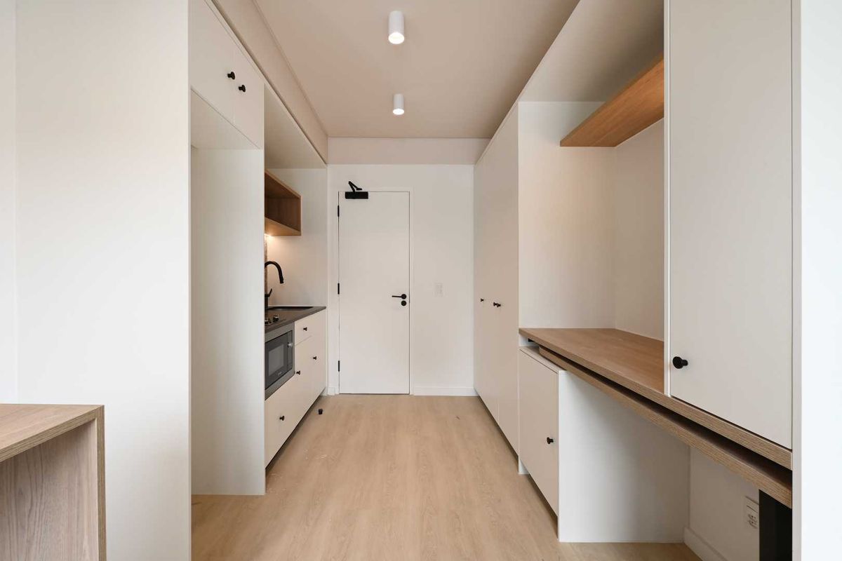 A modern, minimalist kitchen area featuring clean white cabinetry, integrated appliances, light wood-tone flooring, and recessed ceiling lighting in a compact, efficient layout.