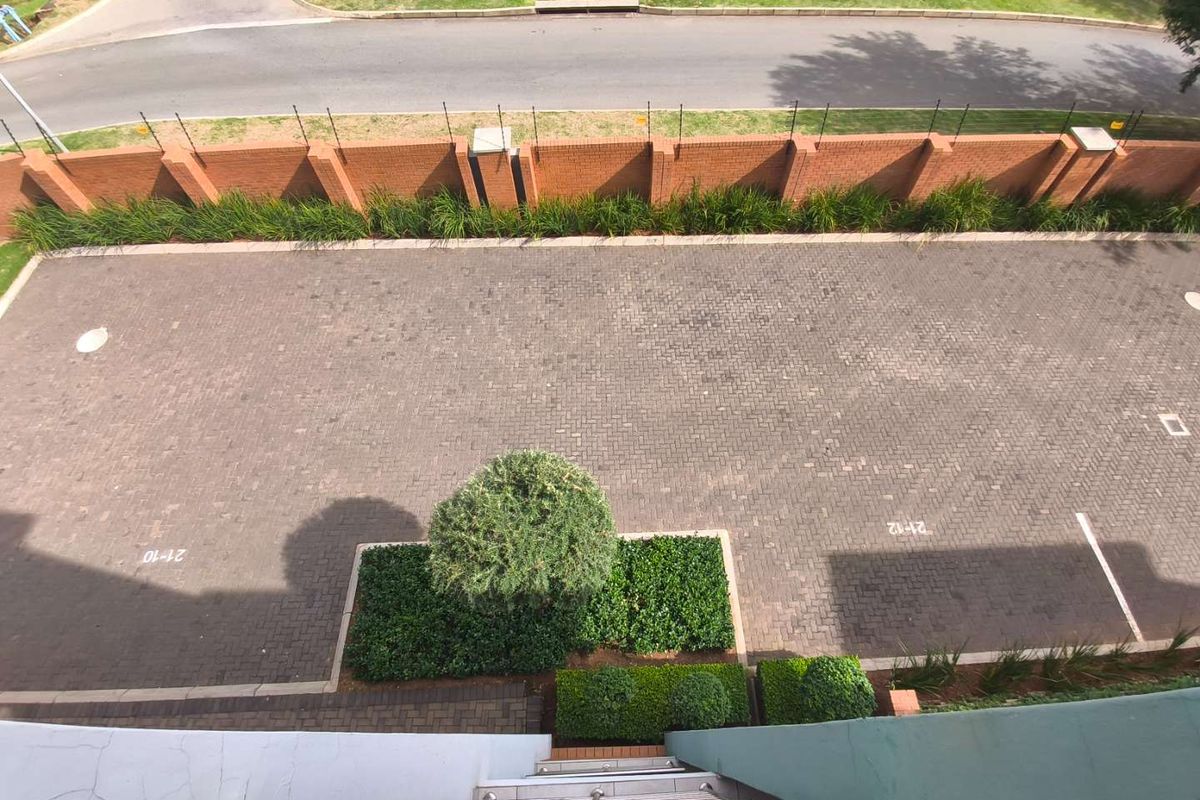Aerial view of the apartment complex parking area featuring paved driveway, landscaped garden beds, and perimeter brick wall with electric fencing.