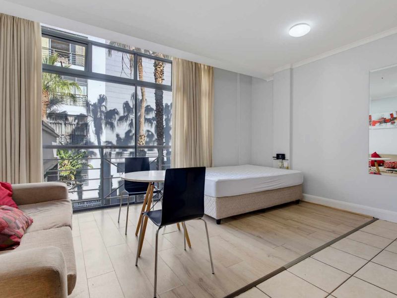 An open-plan studio apartment featuring large floor-to-ceiling windows, a mix of laminate and tiled flooring, and a bright, functional living and sleeping area.