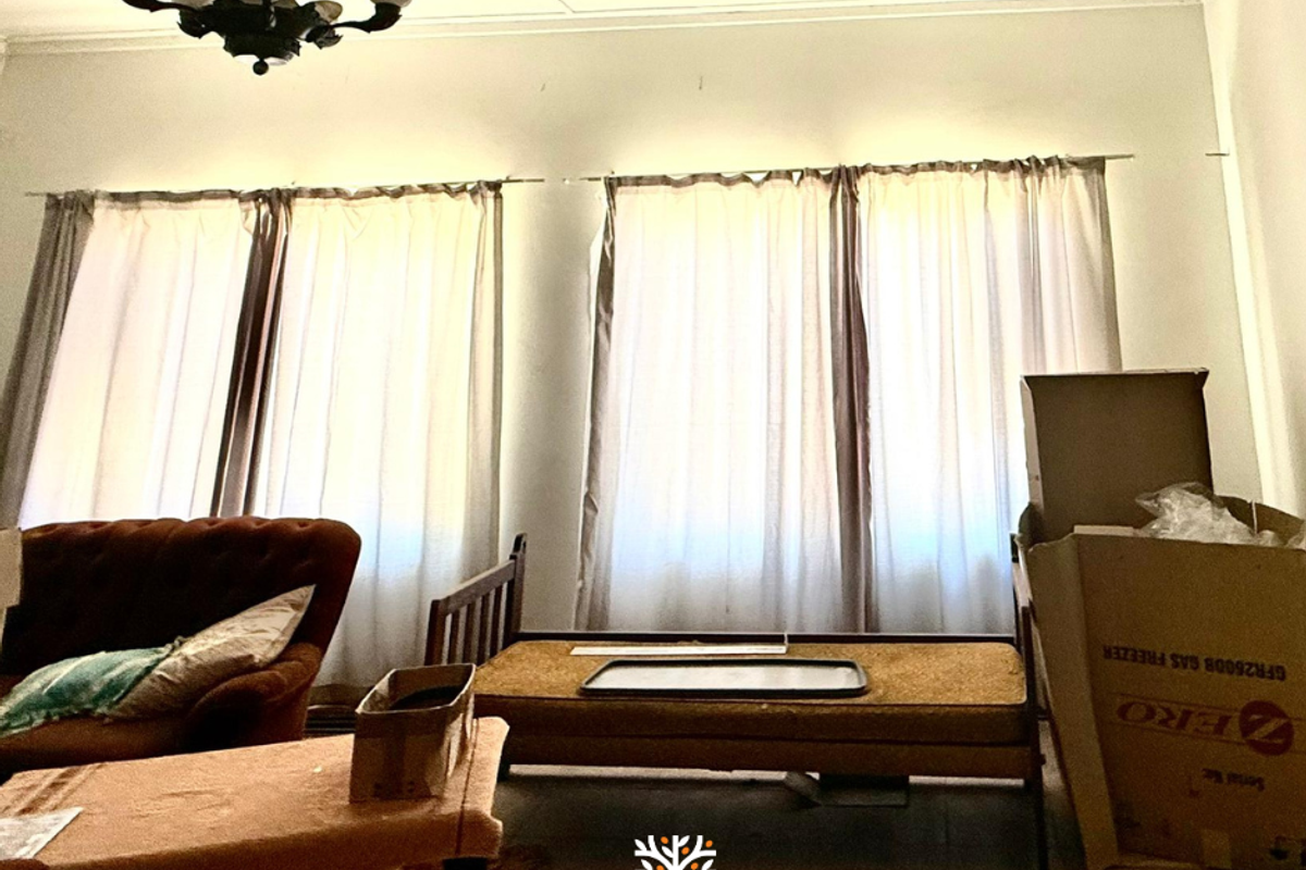 A living area featuring large windows with curtains and a ceiling-mounted light fixture, showing signs of wear and requiring cosmetic updates.