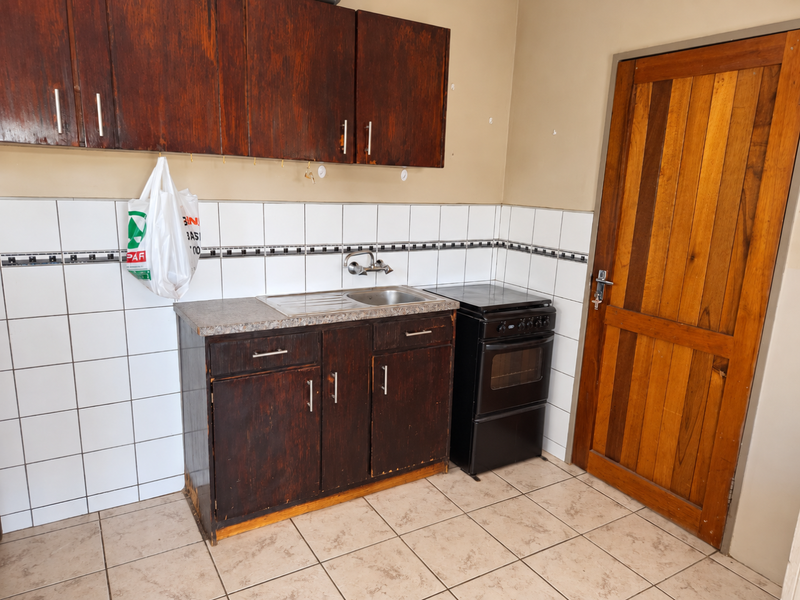 Compact kitchen featuring dark wood cabinetry, tiled backsplash, stainless steel sink, and a freestanding stove, with direct access via a wooden door.