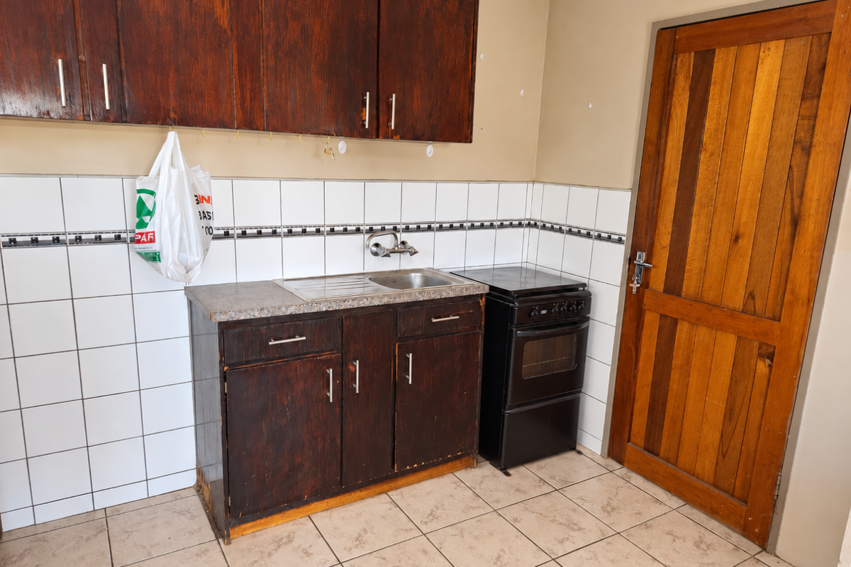Compact kitchen featuring dark wood cabinetry, tiled backsplash, stainless steel sink, and a freestanding stove, with direct access via a wooden door.