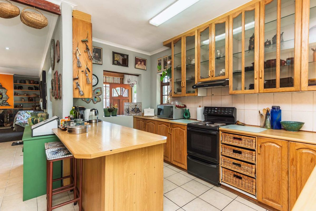 A functional kitchen featuring wooden cabinetry, glass-fronted display cupboards, a tiled backsplash, and a breakfast bar, with a view into the adjacent living area.