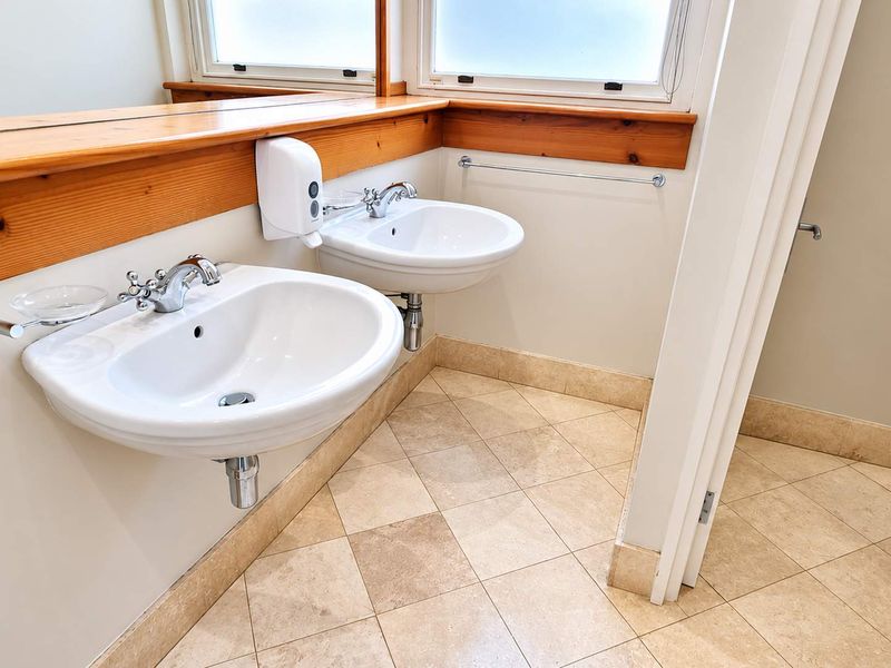 A clean, functional office bathroom featuring dual wall-mounted ceramic basins, a timber vanity shelf, large mirror, and neutral tiled flooring.