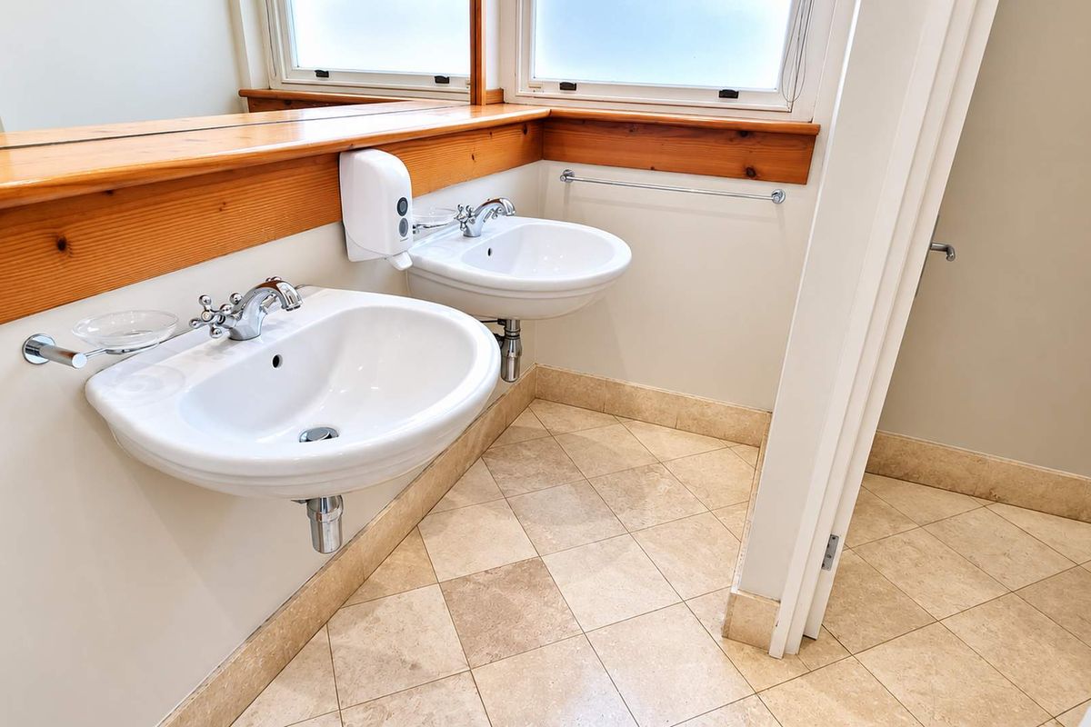 A clean, functional office bathroom featuring dual wall-mounted ceramic basins, a timber vanity shelf, large mirror, and neutral tiled flooring.