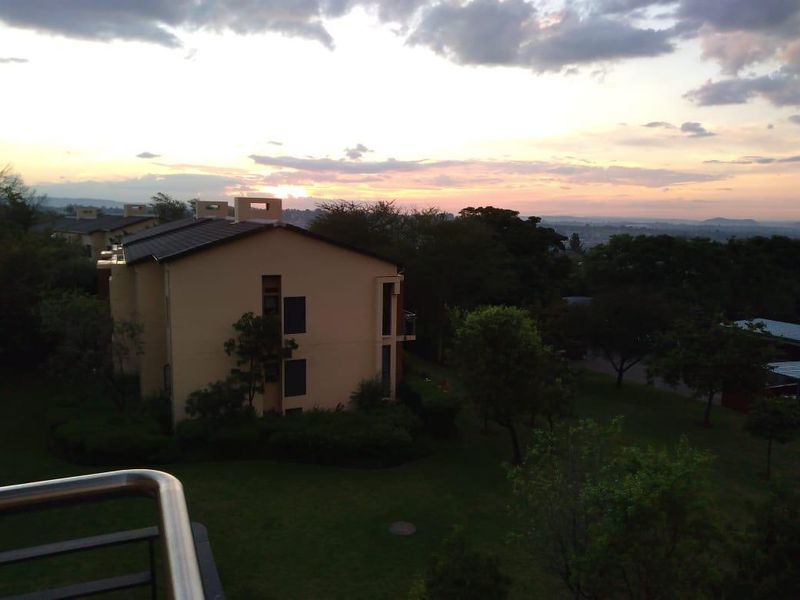 A scenic sunset view overlooking the apartment complex grounds and surrounding landscape, captured from an elevated balcony with a stainless steel railing.