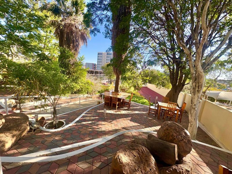 A spacious outdoor patio area featuring patterned paving, mature trees, and a landscaped garden setting, ideal for office break-out space or outdoor meetings.