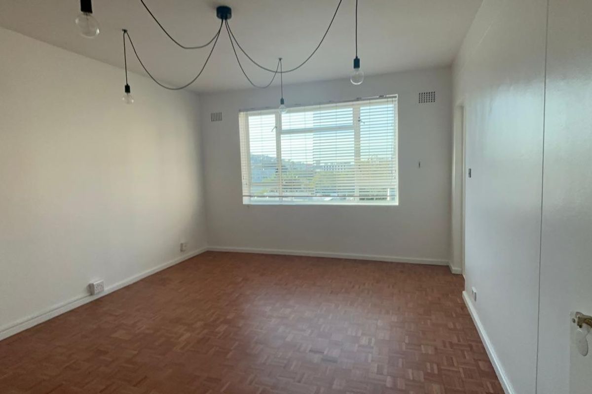 A bright and spacious empty living room featuring classic parquet flooring, large windows with venetian blinds, and neutral white walls.