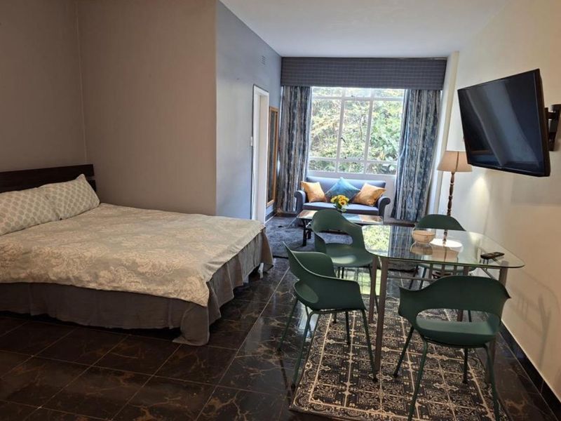 An open-plan studio apartment featuring dark tiled flooring, a wall-mounted television, and a large window providing natural light.