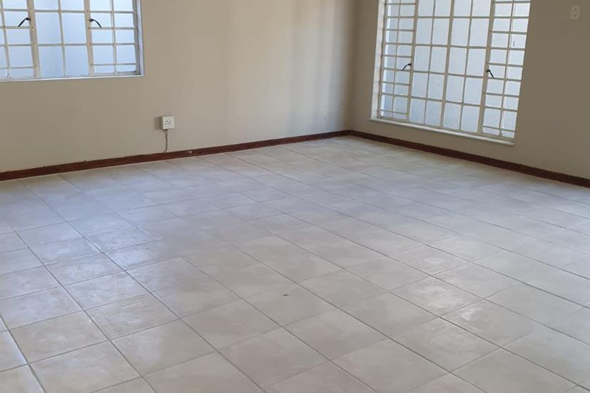 A spacious, empty living area featuring neutral tiled flooring, large windows with security bars, and a simple ceiling-mounted light fixture.
