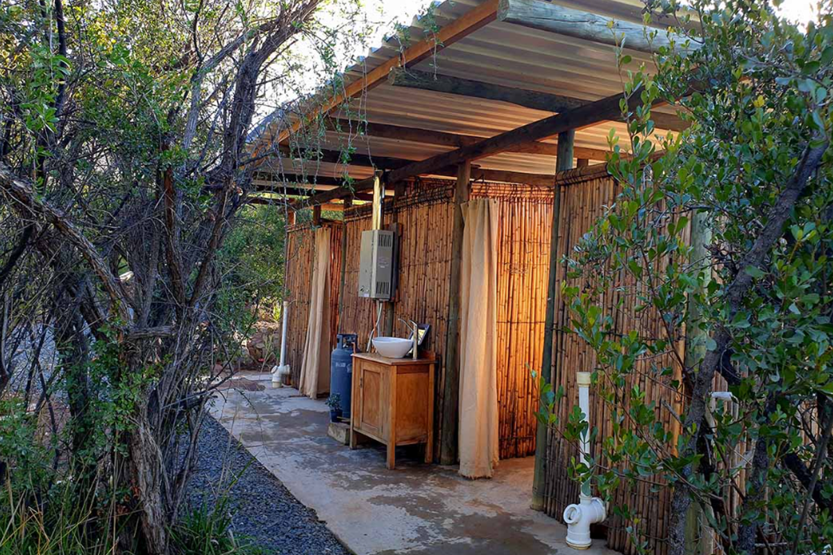 Outdoor bathroom facility featuring a rustic bamboo structure, corrugated roof, and a wall-mounted gas geyser with a vanity basin.