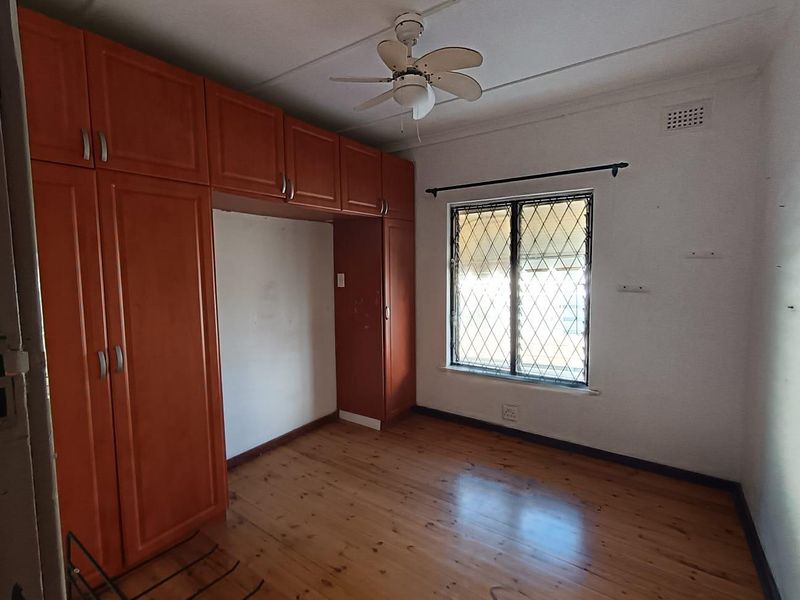 A vacant bedroom featuring wooden flooring, built-in cupboards, and a ceiling fan, with a window fitted with security bars.