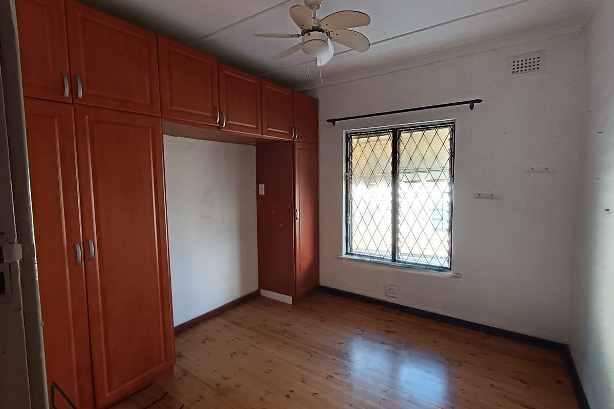 A vacant bedroom featuring wooden flooring, built-in cupboards, and a ceiling fan, with a window fitted with security bars.