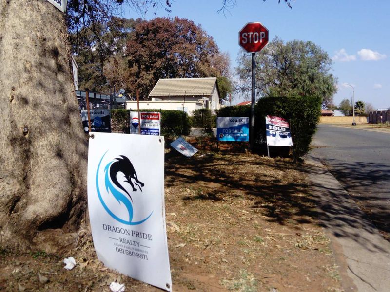 A street-level view showing the neighbourhood approach with multiple real estate signage boards and a mature tree along the roadside.