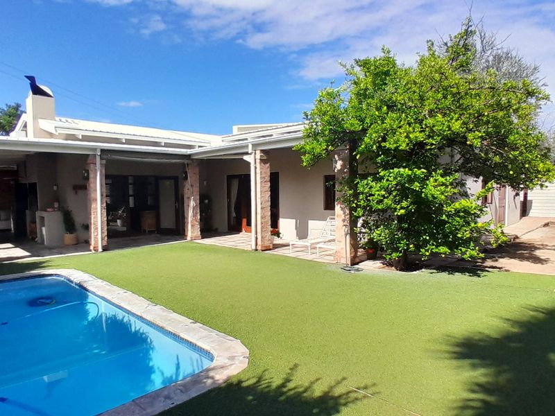 Rear exterior view featuring a swimming pool, artificial lawn, and a covered patio area with brick pillars and an outdoor fireplace.