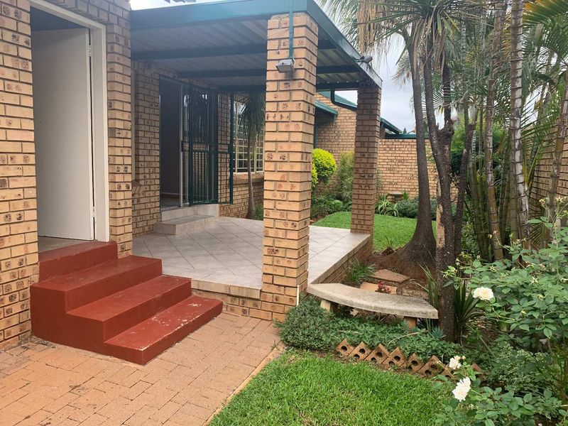 A covered brick patio area with tiled flooring, featuring concrete steps leading to the entrance and a view of the adjacent garden.