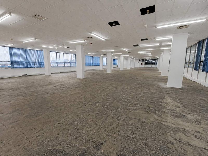 Large, open-plan office space featuring structural columns, suspended ceilings with fluorescent lighting, and expansive windows with vertical blinds.