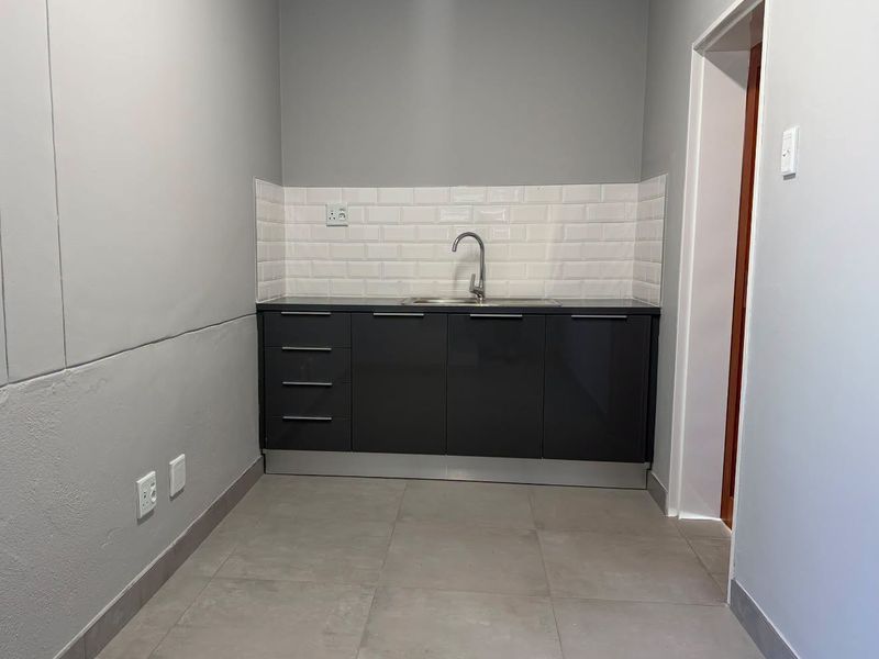 A compact, modern kitchenette featuring dark grey cabinetry, a subway tile backsplash, and a stainless steel sink, finished with neutral large format floor tiles.