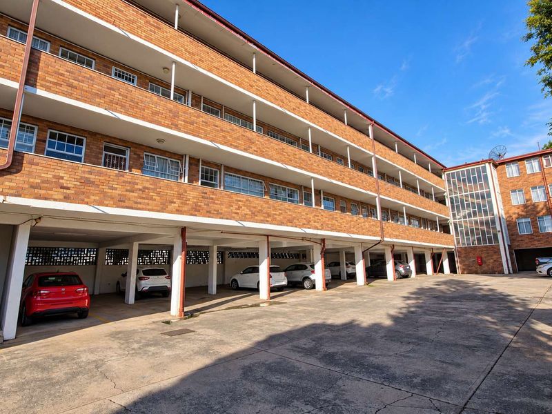 Exterior view of a multi-story brick apartment building featuring covered ground-level parking bays and a concrete driveway.