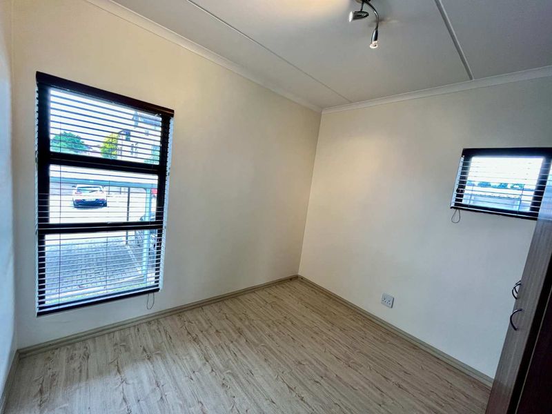 A bright, empty bedroom featuring light-toned laminate flooring, neutral walls, and venetian blinds on the windows.